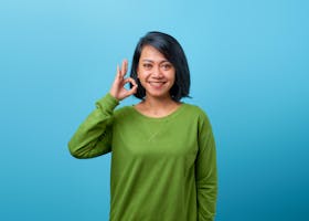 Happy adult woman in green sweater making an okay sign on blue background.