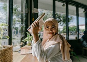 A smiling woman in a hijab reading a book by a window in a cozy cafe in Banda Aceh, Indonesia.